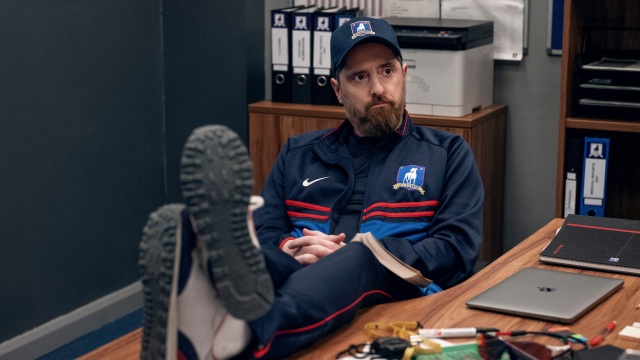 Brendan Hunt as Coach Beard sitting at his desk with feet up