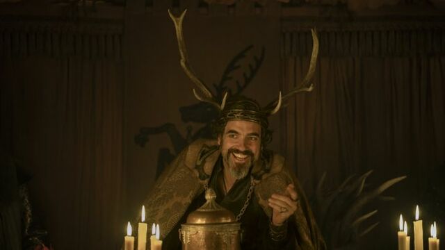 alt="Daniel Ings as Ser Lyonel Baratheon wearing a stag-antler crown and fur cloak, seated at a candlelit feast in HBO’s A Knight of the Seven Kingdoms."