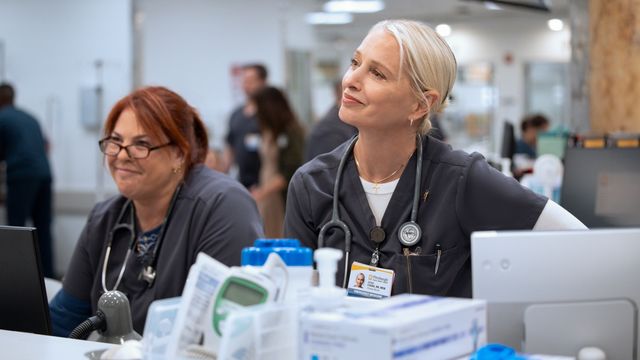 alt="Dana Evans sits at a hospital station, smirking while listening to someone off-camera."