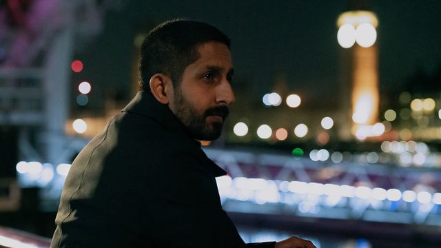 alt="Sagar Radia as Rishi Ramdani leaning on a bridge railing at night, city lights glowing behind him."