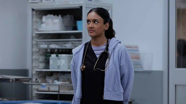 alt="Victoria Javadi stands in a hospital room wearing scrubs and a hoodie, listening attentively."